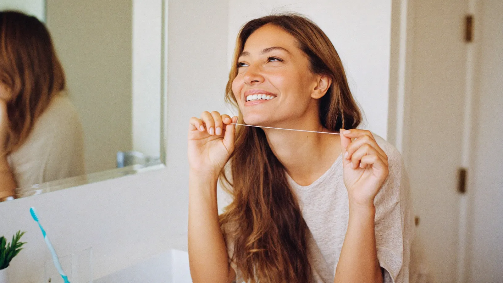 A smiling woman holding a strand of dental floss in a bright bathroom, with a toothbrush on the counter beside her.