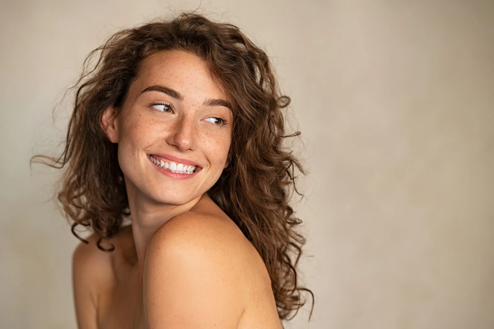 A woman with freckles and curly hair glancing over her shoulder mid-laugh, her natural smile unretouched, the kind of result cosmetic dentistry aims to enhance rather than replace.