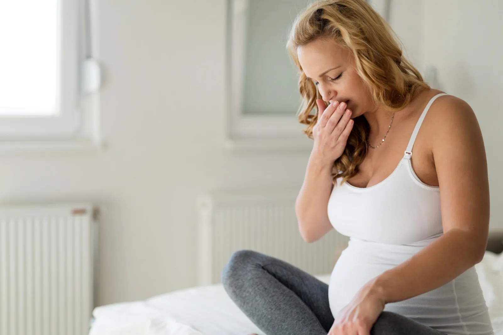 A pregnant woman sitting on the edge of a bed with a hand raised to her mouth, one of the life stages when shifting hormone levels can change how the gums and teeth respond.