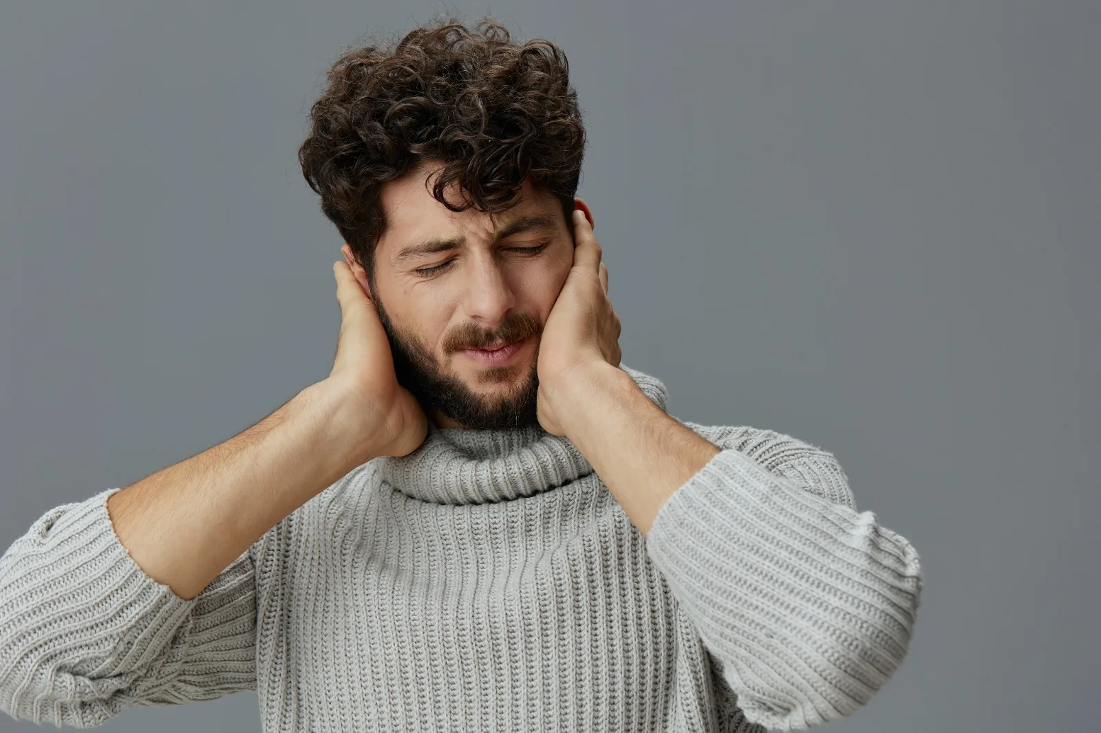 A man with his eyes closed pressing both hands against his jaw and ears, the overlapping discomfort of jaw tension, headache and ear ringing that often turns out to share one cause.