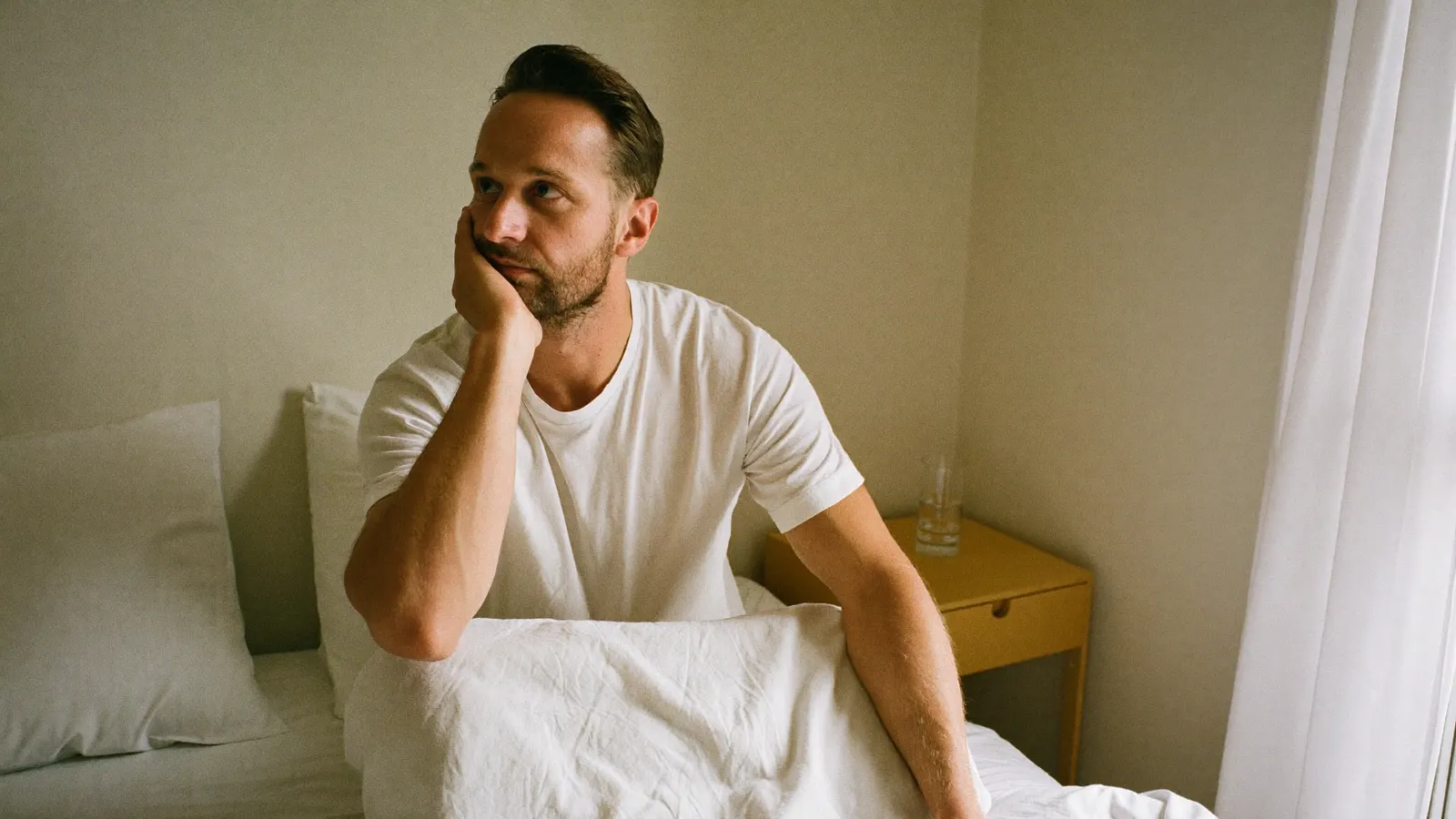 A tired man sitting up in bed in the morning with his hand pressed to his jaw, a common sign of nighttime teeth grinding.